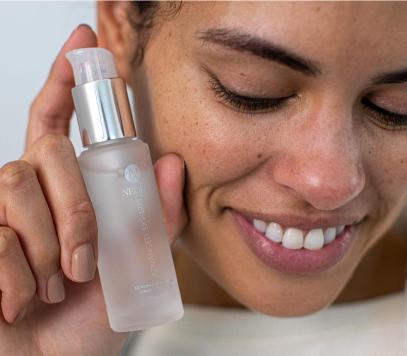 Woman holding Neora’s Advanced SIG-1273 Concentrated Serum 
                        in her hand next to her face while smiling 